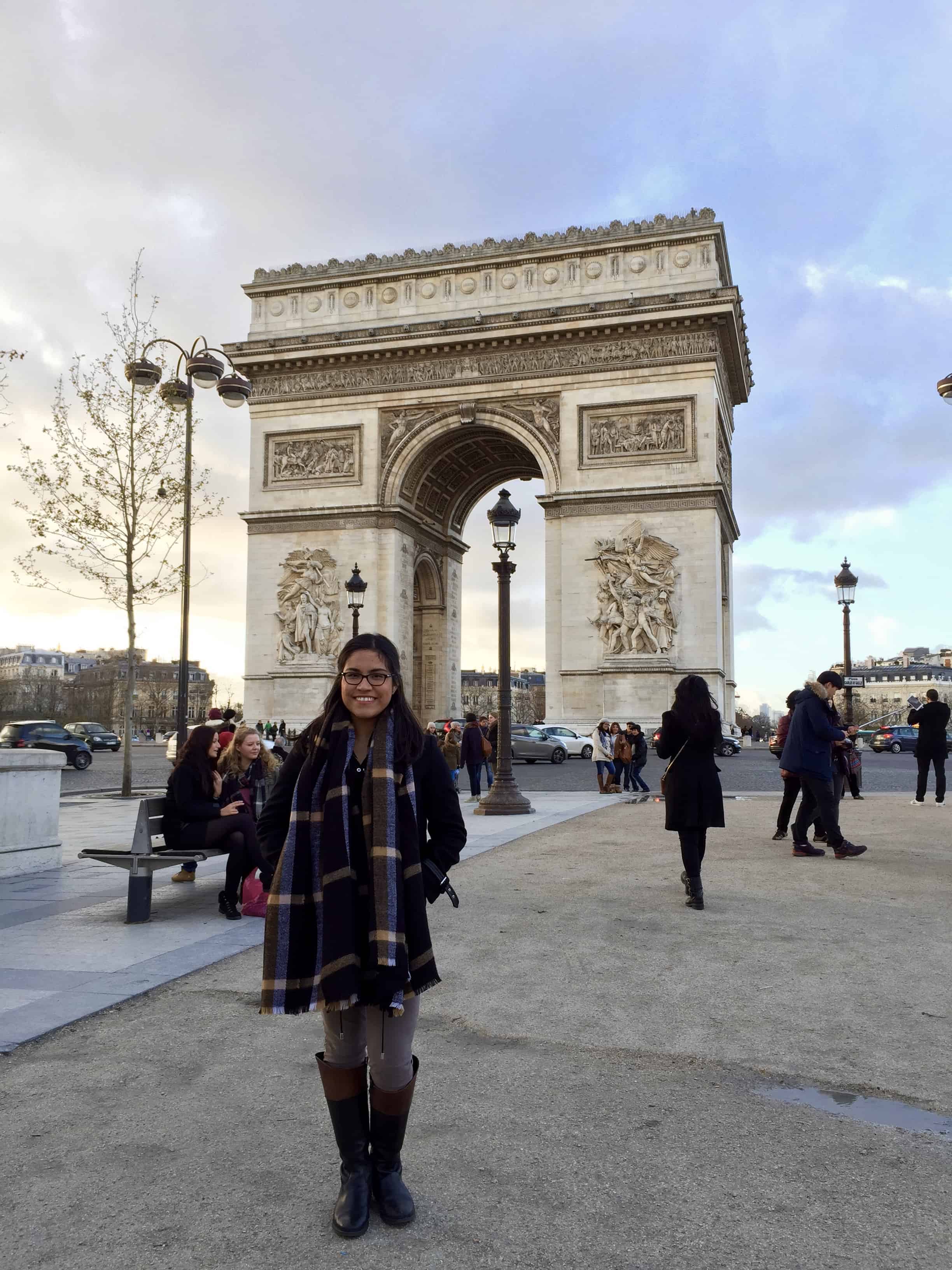 girl with glasses and black hair and scarf standing in front of the arc de triumph