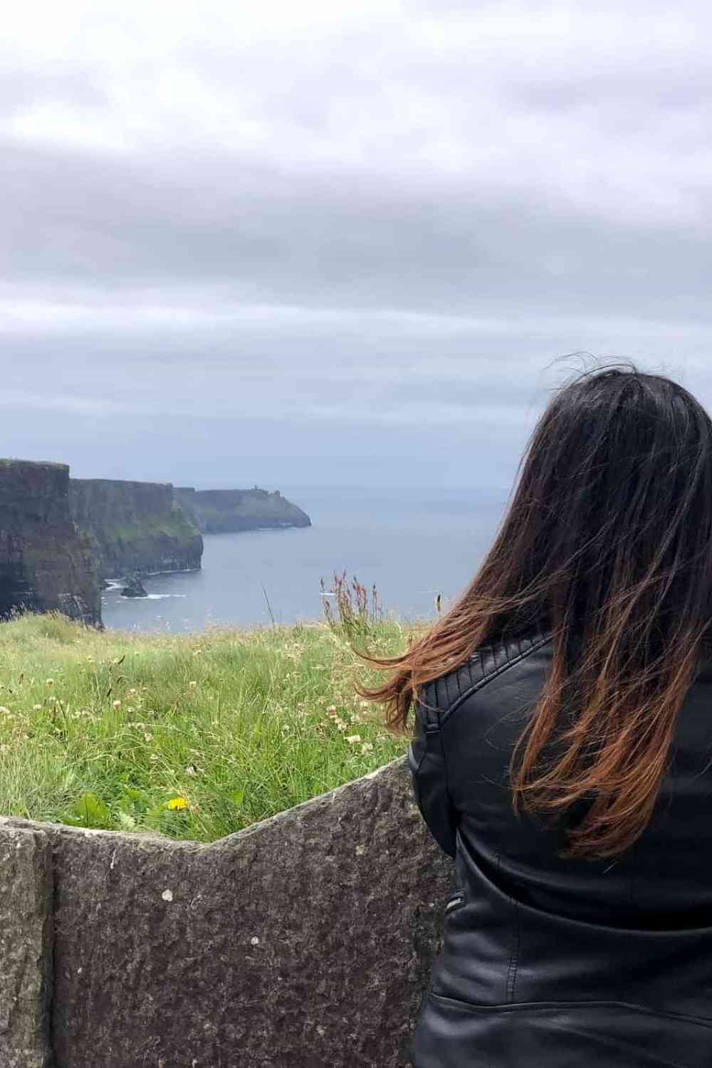 girl with brown hair and black leather jacket looking out onto the sea and mountain cliffs at the cliffs of moher, ireland
