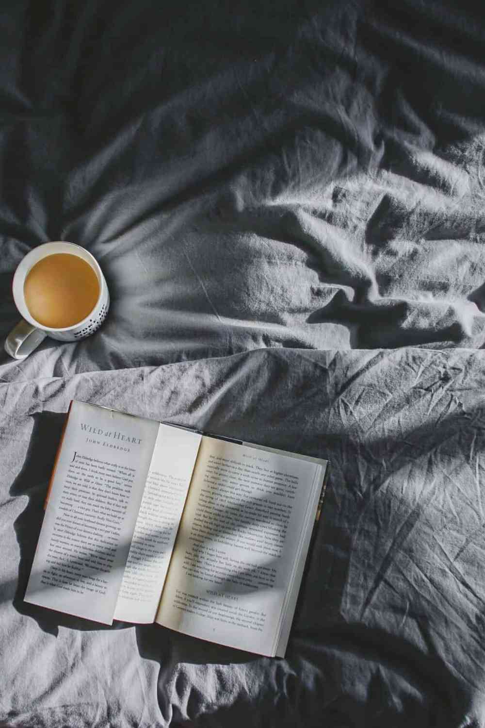 a cup of coffee on a bed with gray sheets and an open book with the sunlight
