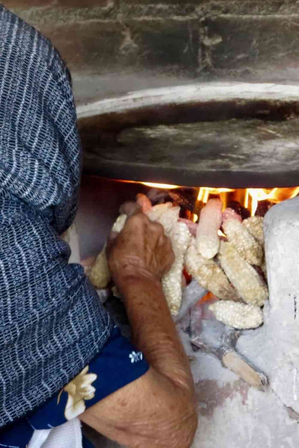 an older woman with a blue headscarf putting dried corn inside a fire stove to make tortillas