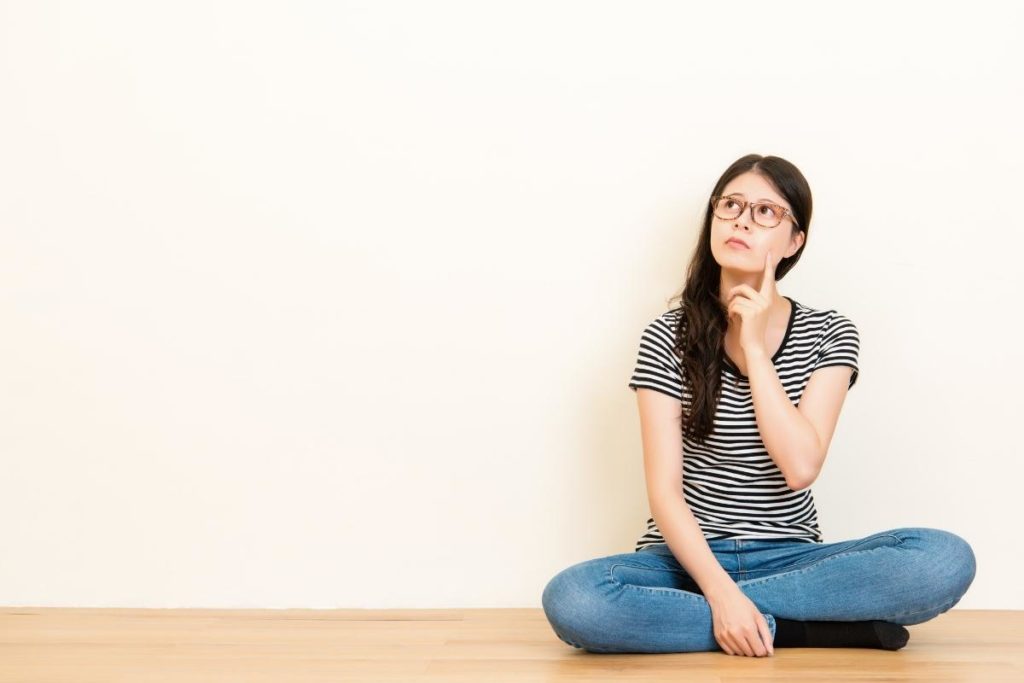 girl sitting down with glasses with a finger up to her cheek thinking and looking away