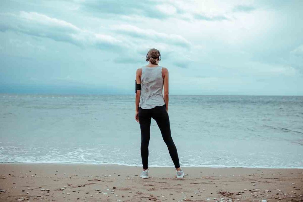 girl with workout tank top and black leggings standing in front of ocean while listening to music on her workout