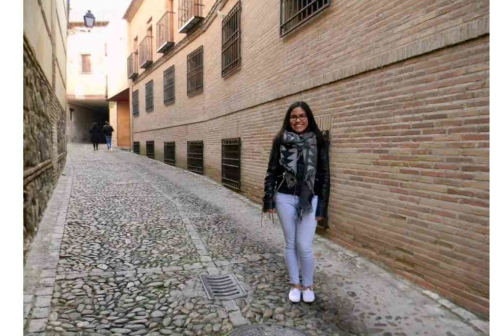 girl with black hair and a black leather jacket and glasses standing in European cobblestone streets 
