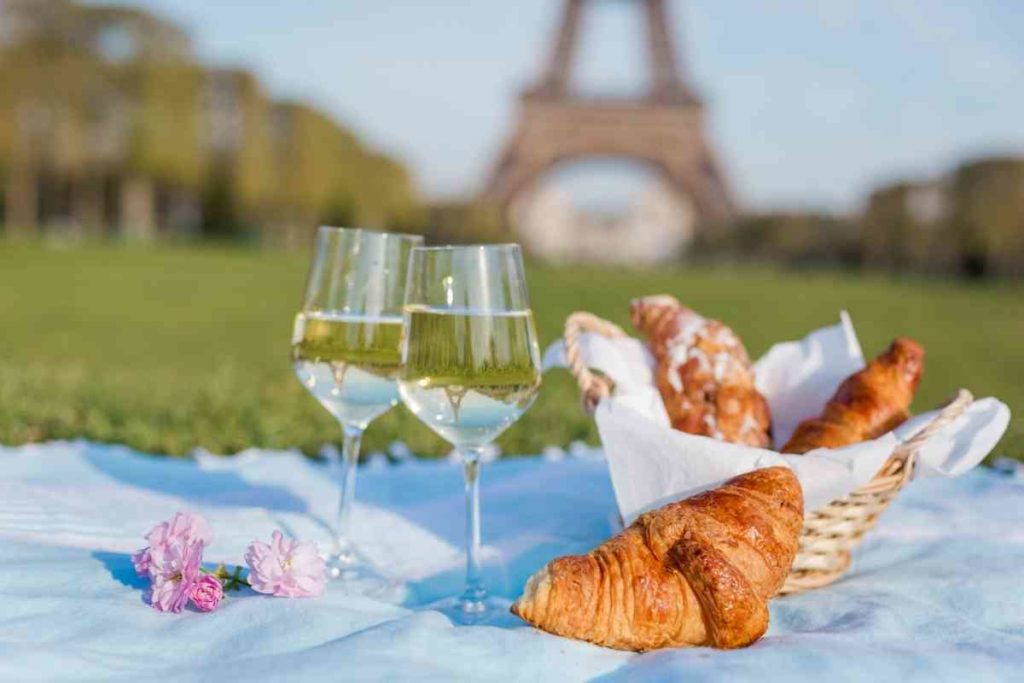 A close up of 2 glasses of wine with a basket of croissants in a picnic in front of the Eiffel Tower in Paris on a sunny day