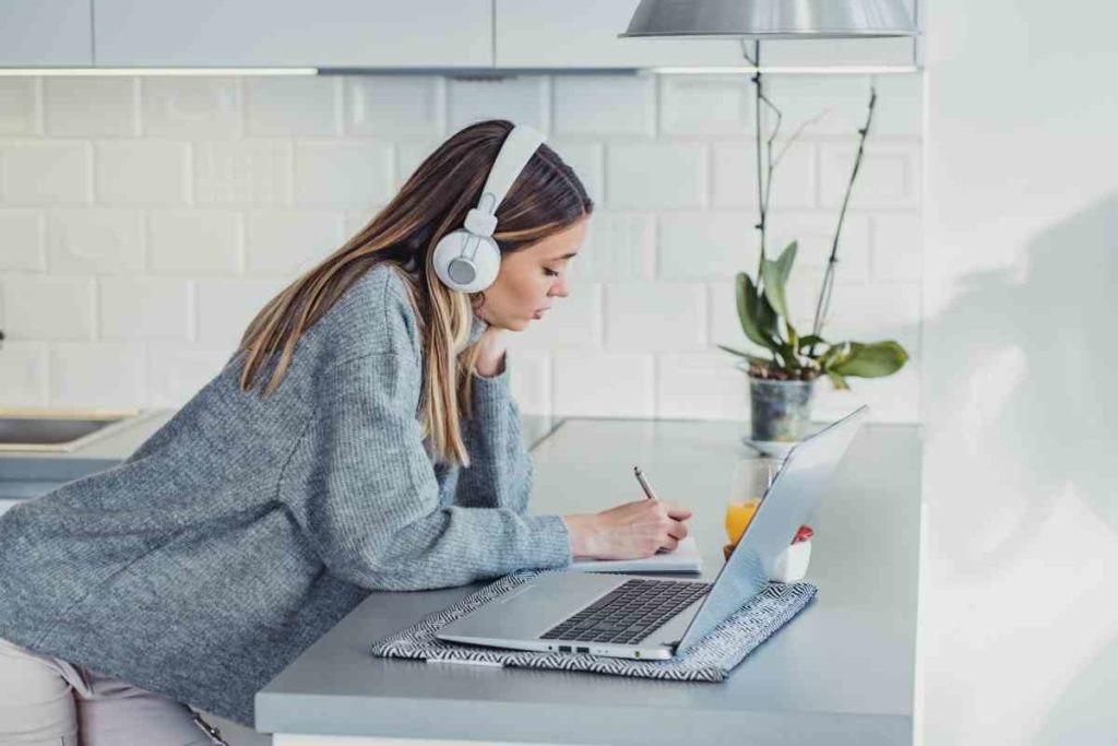 girl doing homework and attending class online with her laptop and headphones taking notes in her kitchen