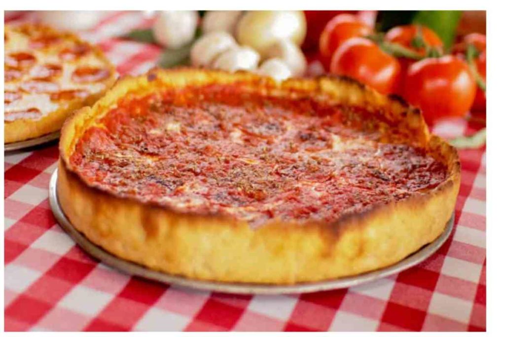 close up of Chicago style deep dish pizza with tomatoes and onions in the background and a red and white checkered mantle