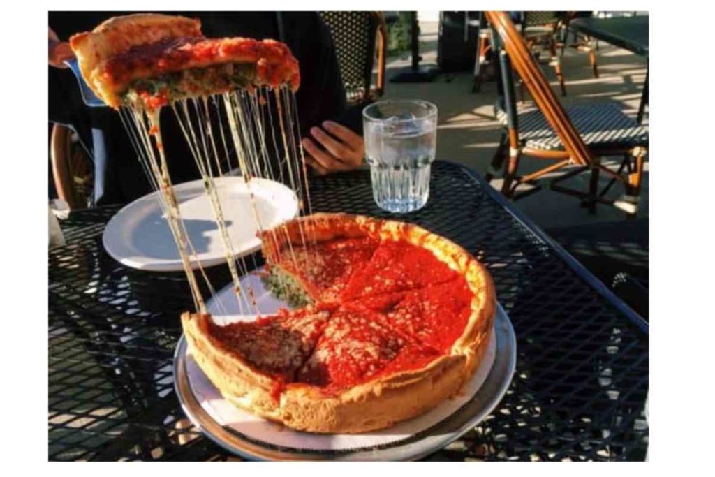 A slice of cheesy deep-dish pizza on a black table with a cup of water and white plate next to it