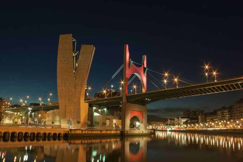 The La Salve Bridge, a stunning spectacle on the Bilbao Free Tours, gleams with red and pink tones against the evening sky, mirrored on the Nervión River's surface.