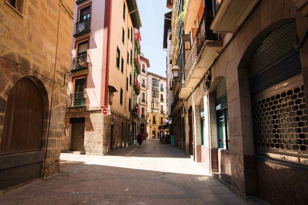 On a walking tour in Bilbao, explore the Old Town's cobblestone paths flanked by buildings painted in a palette of reds, yellows, and creams, adorned with green balcony plants on a sunny day.