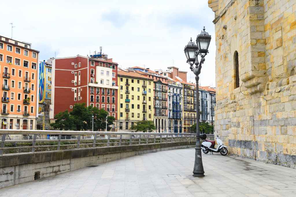 A quiet Bilbao free walking tour moment by the Nervión River, where a lone white scooter rests beside a promenade with golden stone walls and a mosaic of colorful buildings.