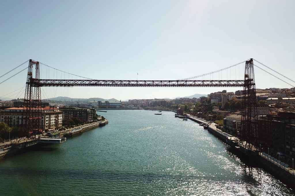 The Vizcaya Bridge, captured on a tour in Bilbao, stretches across the blue river, its red framework contrasting with the green landscape under a clear azure sky in Getxo, only 20 minutes from Bilbao.