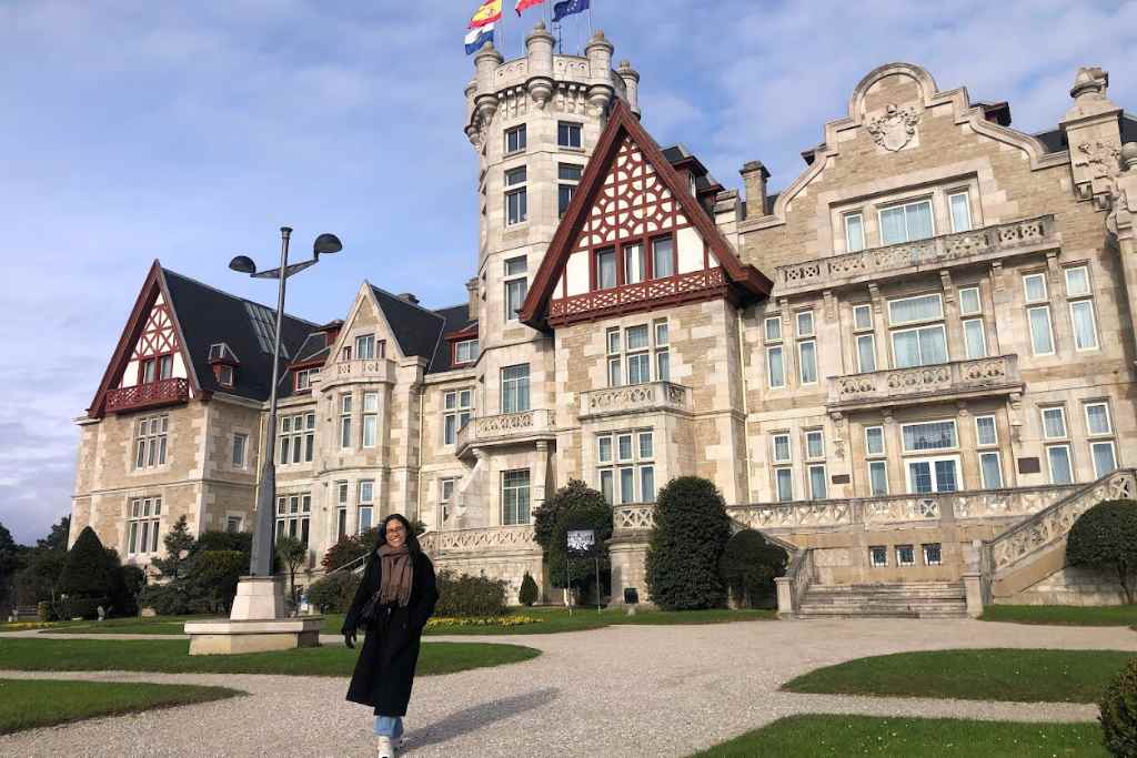A joyful girl wearing a long black coat with glasses and dark hair stands before the grand Palacio de la Magdalena in Santander, exemplifying 'why visit Santander' with its stunning architectural beauty and royal history.