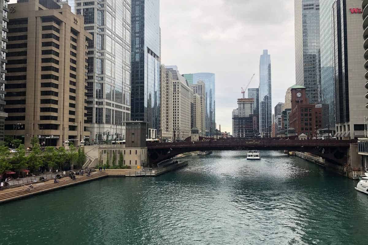 View along the Chicago River showcasing a vibrant mix of modern and historical architecture, with skyscrapers lining both sides of the waterway. A pedestrian bridge, busy with walkers, connects the city sides, under which tour boats and water taxis navigate the river. This urban riverscape highlights safe and scenic routes for solo travelers, providing a peaceful yet bustling atmosphere, perfect for exploring Chicago's architectural marvels and city life alone.