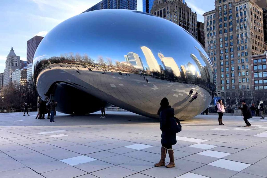 A solitary traveler stands admiring the iconic Cloud Gate sculpture, also known as 'The Bean', in Chicago's Millennium Park. The sculpture's shiny, mirror-like surface reflects the clear blue sky and surrounding skyscrapers. This scene illustrates a popular and safe destination for solo travelers in Chicago, showcasing a must-visit spot that allows visitors to appreciate the city's architecture and vibrant urban atmosphere alone.