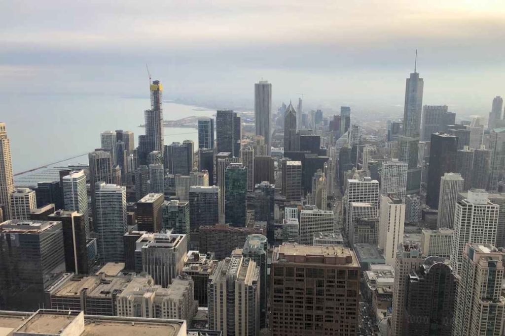 Aerial view of downtown Chicago featuring a dense cluster of skyscrapers extending towards Lake Michigan, captured from a high vantage point. Prominent buildings pierce the skyline under a hazy sky, illustrating a bustling urban landscape. This image exemplifies the vast cityscape solo travelers can explore, emphasizing the diverse architectural experiences and safe, engaging environment Chicago offers to visitors, especially female travelers exploring the city alone.