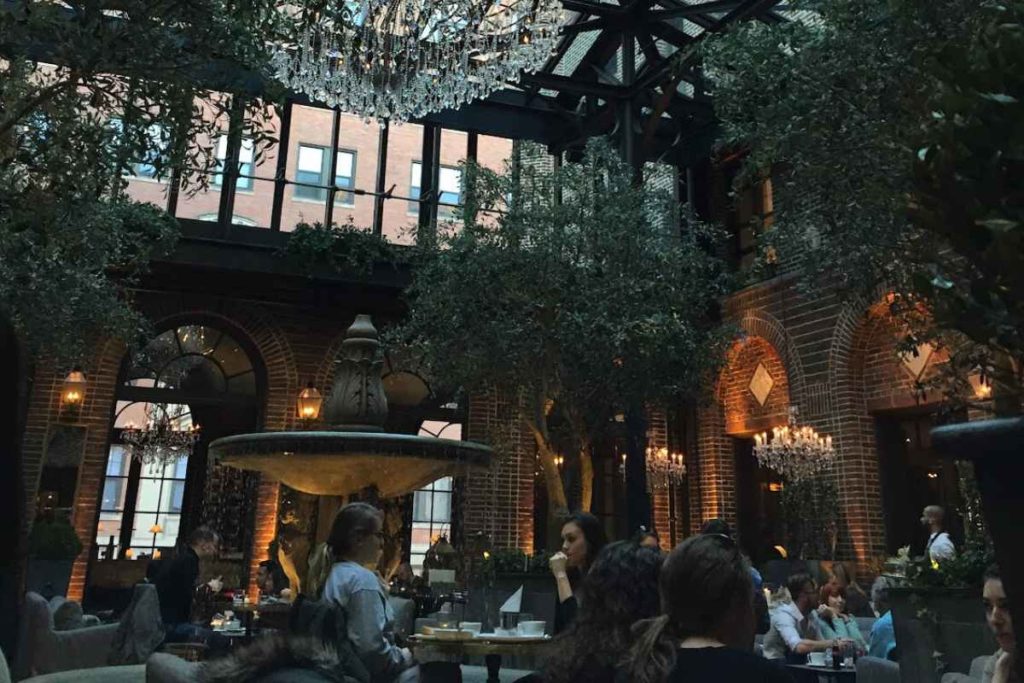 A bustling indoor restaurant scene in Chicago, showing solo and group diners in a relaxed setting with elegant décor. The venue features a large fountain as the centerpiece, surrounded by lush greenery and exposed brick walls, under a glass roof and crystal chandeliers. This image captures the vibrant dining experience and architectural beauty, suitable for exploring things to do in Chicago alone, illustrating why the city is safe and welcoming for female travelers.