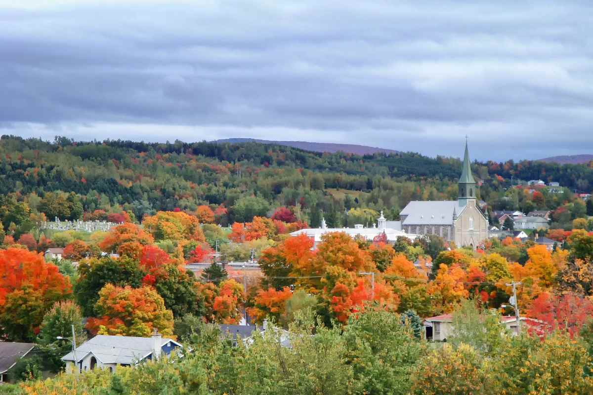 A scenic view of a small Quebec town surrounded by vibrant fall foliage and forested hills, perfect for autumn day trips from Montreal.