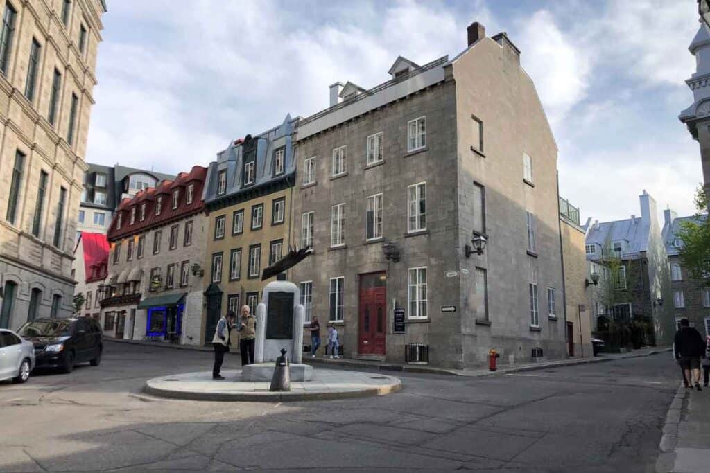 A quiet street corner in Old Québec City featuring historic stone and colorful row houses with steep roofs, a small circular monument in the center with people gathered around it, and a couple walking down the street in the background.