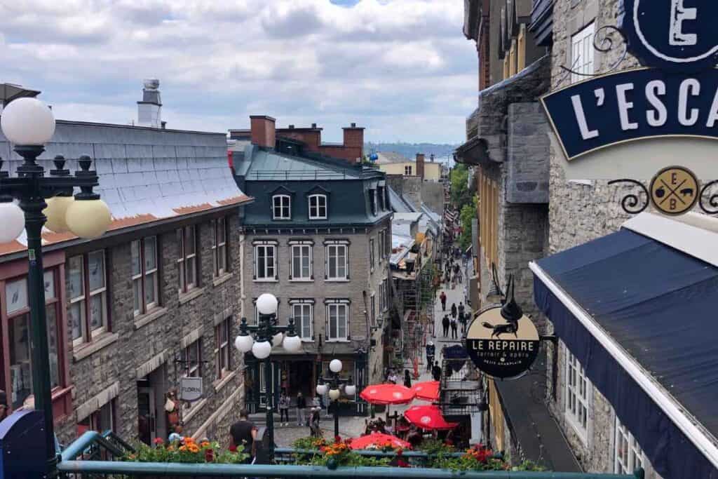 A scenic view of a narrow cobblestone street in Old Québec City, lined with historic stone buildings and vibrant signage. Red patio umbrellas and outdoor diners are visible at the bottom of the slope, including signs for restaurants like "Le Repaire" and "L’ESCAPADE." The street is bustling with pedestrians, while flower boxes and classic street lamps add charm to the scene. The overcast sky and layered rooftops create a cozy, European village feel.