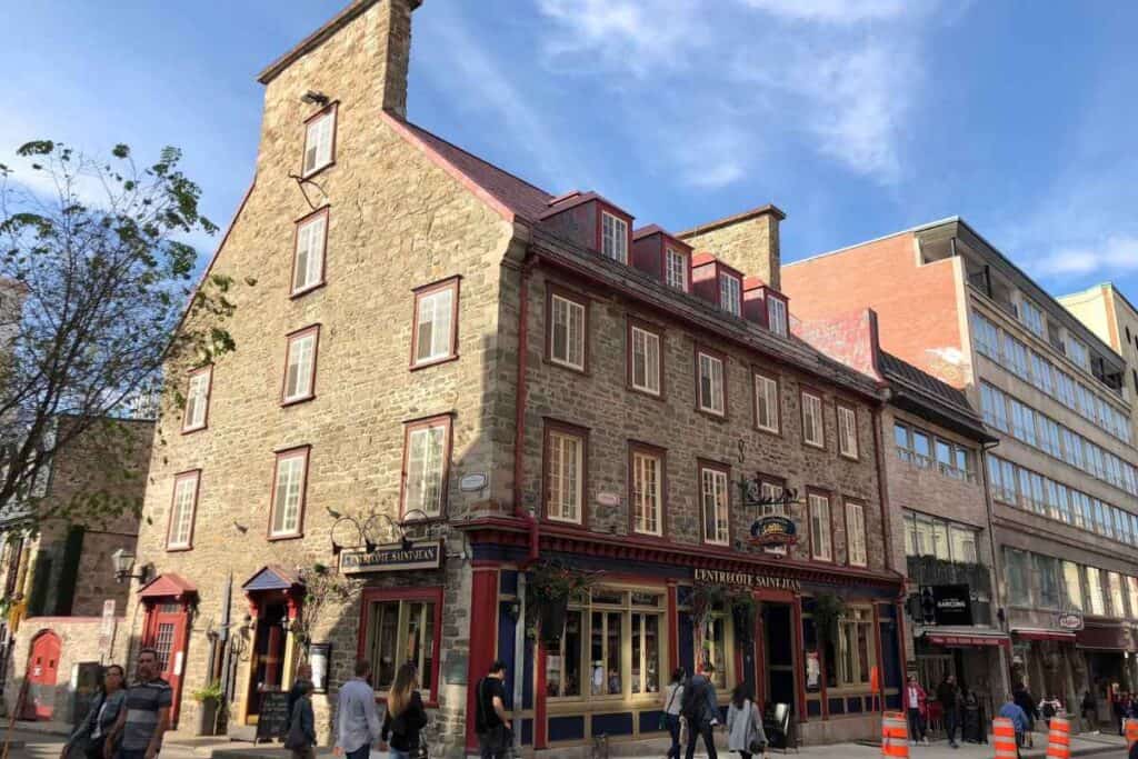A historic stone building in Old Québec City with red trim and a slanted roof, housing the French restaurant “L’Entrecôte Saint-Jean.” The charming facade includes hanging plants, vintage-style signage, and multiple windows. People stroll by on the sidewalk, and a few orange traffic cones line the edge of the street. A mix of old and modern buildings is visible in the background under a blue sky.