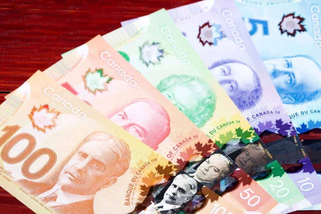 A fanned-out arrangement of colorful Canadian banknotes in denominations of 100, 50, 20, 10, and 5 dollars, each featuring prominent Canadian historical figures and a transparent maple leaf security feature, displayed on a red wooden surface.