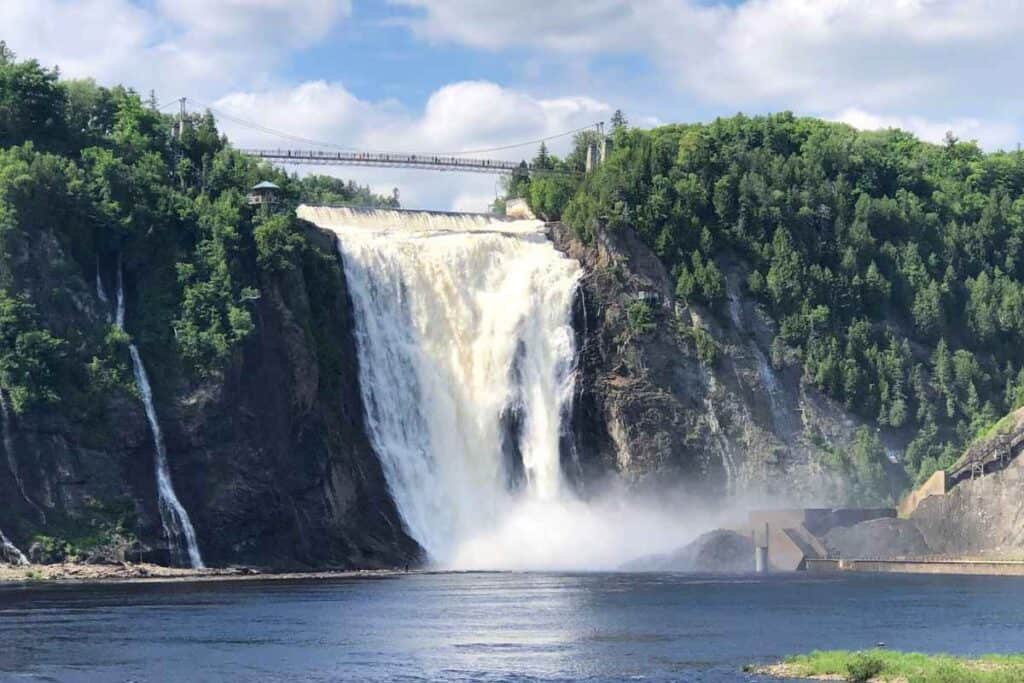 A wide view of Montmorency Falls cascading powerfully down a rocky cliff surrounded by lush green trees, with mist rising at the base and a suspension bridge stretching across the top of the falls under a partly cloudy sky.
