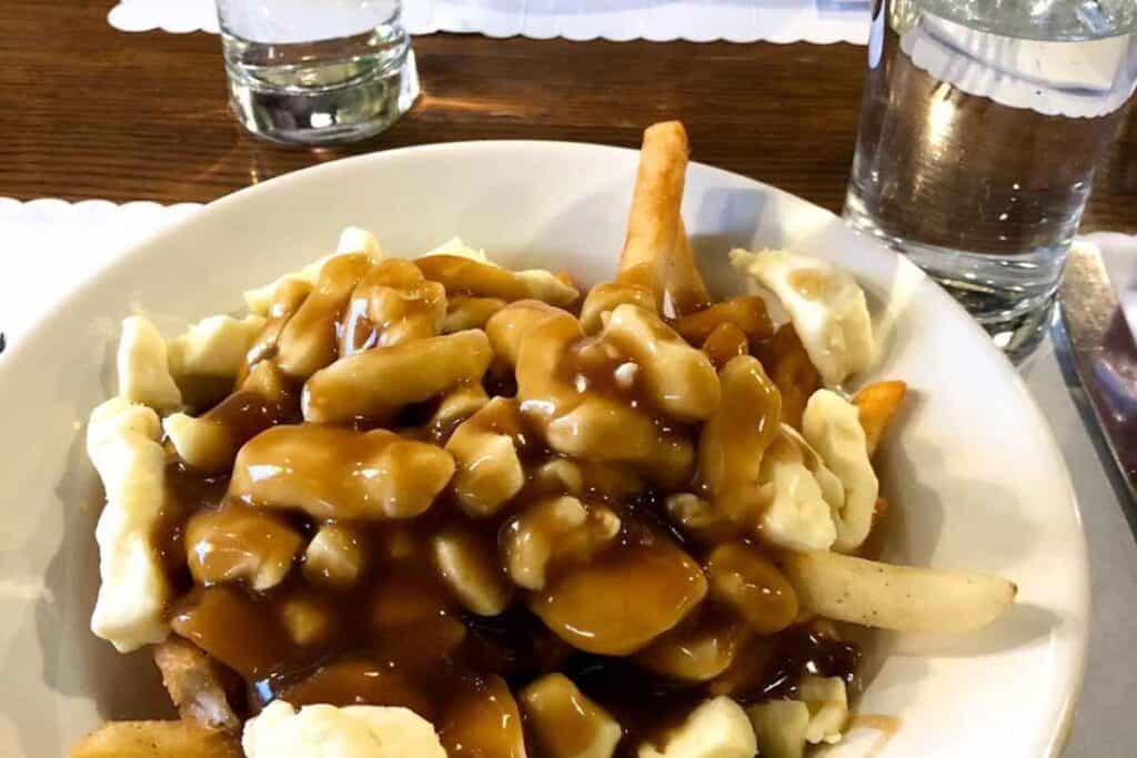 Close-up of a bowl of classic poutine, a Canadian dish made with French fries topped with cheese curds and hot brown gravy. The bowl is placed on a wooden table, with two glasses of water in the background.
