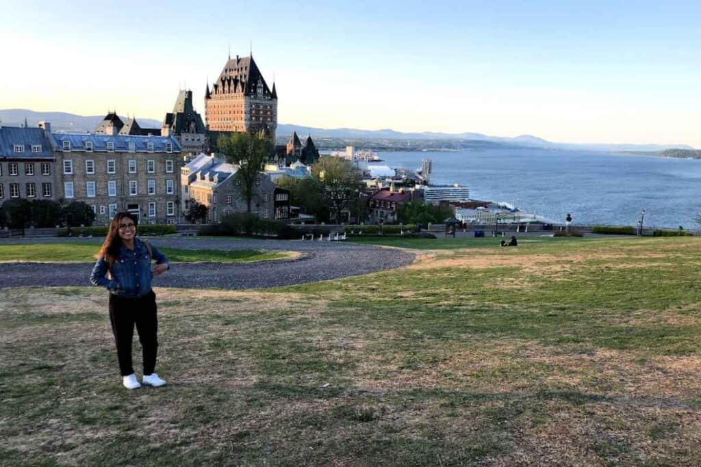 Woman standing on a grassy hill in front of the historic Château Frontenac in Quebec City, Canada, with the Saint Lawrence River and a docked cruise ship in the background.