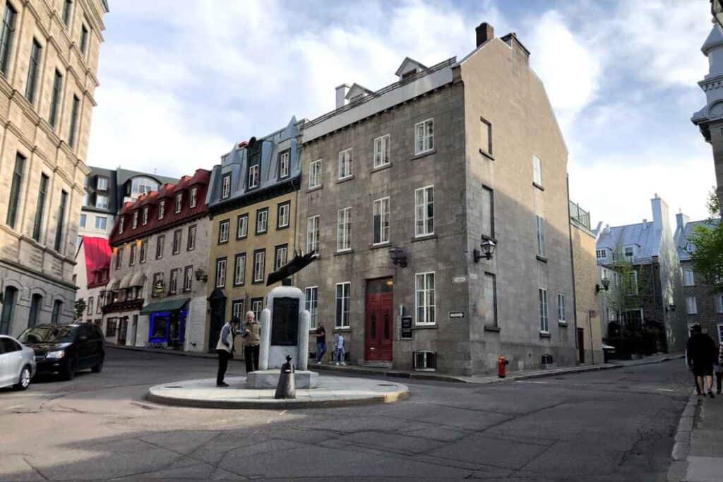 Street corner in Old Quebec with historic stone buildings, colorful facades, and a small monument in the center where two people are reading an inscription.