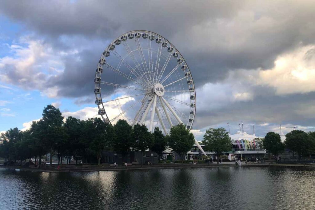 La Grande Roue de Montréal ferris wheel stands tall against a moody sky, reflecting off the water in the Old Port at sunset.