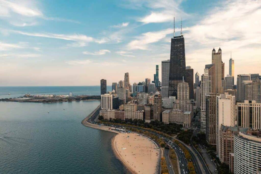 Aerial view of downtown Chicago’s skyline with the John Hancock Center towering over the city, a sandy beach curving along Lake Michigan, and Navy Pier visible in the distance under a partly cloudy sky.